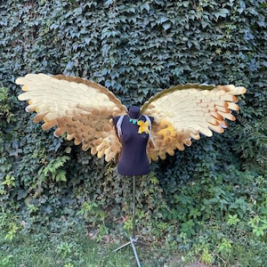 Op de afbeelding: Goudkleurige kostuumvleugels met een gelaagd veermotief, tentoongesteld op een zwarte mannequin. De vleugels zijn gedetailleerd met tinten goud en bruin, en de mannequin is versierd met een ketting en een gele bloem. De achtergrond heeft weelderig groen gebladerte.