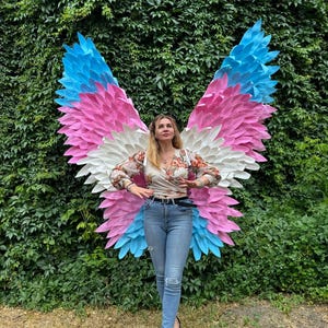 May include: A person standing in front of a wall of green foliage, wearing a floral top, blue jeans, and black heels. Large, colorful butterfly wings in shades of blue, pink, and white are behind the person.