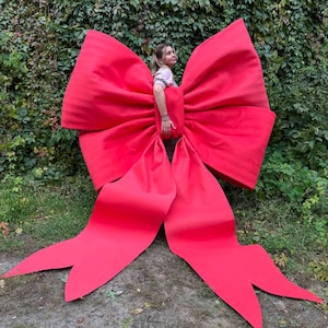Pode incluir: Um grande laço vermelho brilhante, com longas caudas esvoaçantes, está em pé em uma área gramada. O laço é feito de um material liso, possivelmente cetim. O laço é grande o suficiente para que uma pessoa fique dentro dele.