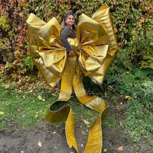 May include: A large, golden bow with long, flowing ribbons. The bow is made of a shiny, textured material and is positioned in front of a backdrop of green and red foliage. The bow is large enough for a person to stand inside.