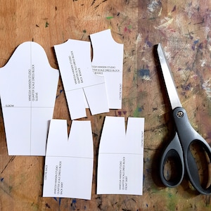 Könnte beinhalten: Ein Satz von fünf weißen Papier-Kleidermustern für ein Kleid-Mieder, einen Rock und einen Ärmel. Die Muster sind mit dem Text "Vanessa Hansen Studio Quarter Scale Dress Block" und dem entsprechenden Teil des Kleides beschriftet. Eine silberne Schere liegt auf einer Holzoberfläche.