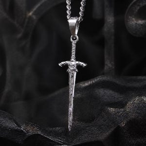 May include: A silver-toned metal pendant shaped like a sword with a textured, weathered finish, suspended from a matching silver-toned rope chain. The sword features a detailed crossguard and a pointed blade, hanging vertically as a necklace.