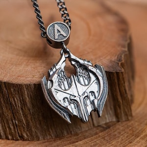 May include: A silver-colored pendant necklace with a detailed, geometric design. The pendant hangs from a chain and features an initial "A" at the top. The necklace is displayed on a wooden surface, showcasing the intricate metalwork.