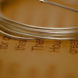Può includere: Primo piano di un filo d'argento avvolto su se stesso, creando una forma circolare. Il filo è su una superficie marrone con il testo "(on for this The How great is so" stampato su di essa.