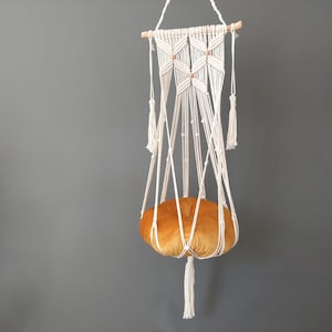 May include: A white macrame hanging cat bed with a mustard yellow velvet cushion. The bed is suspended from a wooden dowel with white tassels.