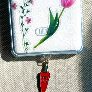 May include: A red chili pepper charm with the words "extra spicy" written on it. The charm is attached to a clear phone case with a floral design.