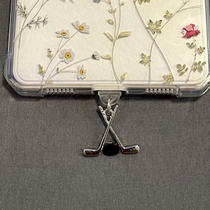 May include: A silver charm featuring two crossed hockey sticks and a black puck. The charm is attached to a phone case with a floral design.