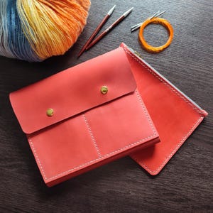 May include: A coral leather knitting needle case and a matching zippered pouch. The case is secured with two gold-coloured studs. A ball of yarn in blue, yellow, and orange, plus knitting needles and a circular needle, are also visible.