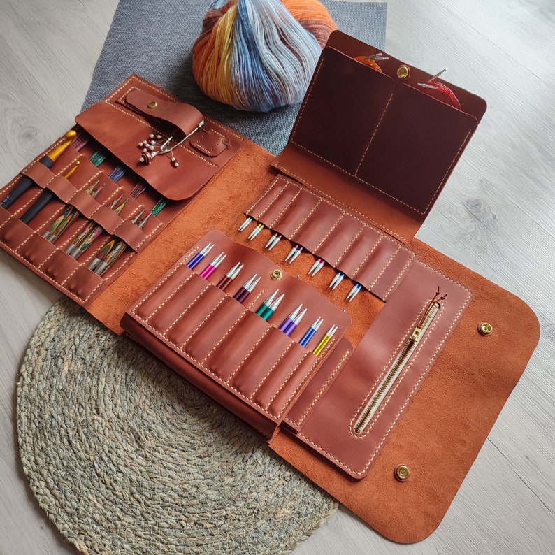 May include: A brown leather knitting needle case with multiple compartments for storing needles. The case is open and the needles are visible. The case has a zipper closure and a flap with a decorative metal buckle.