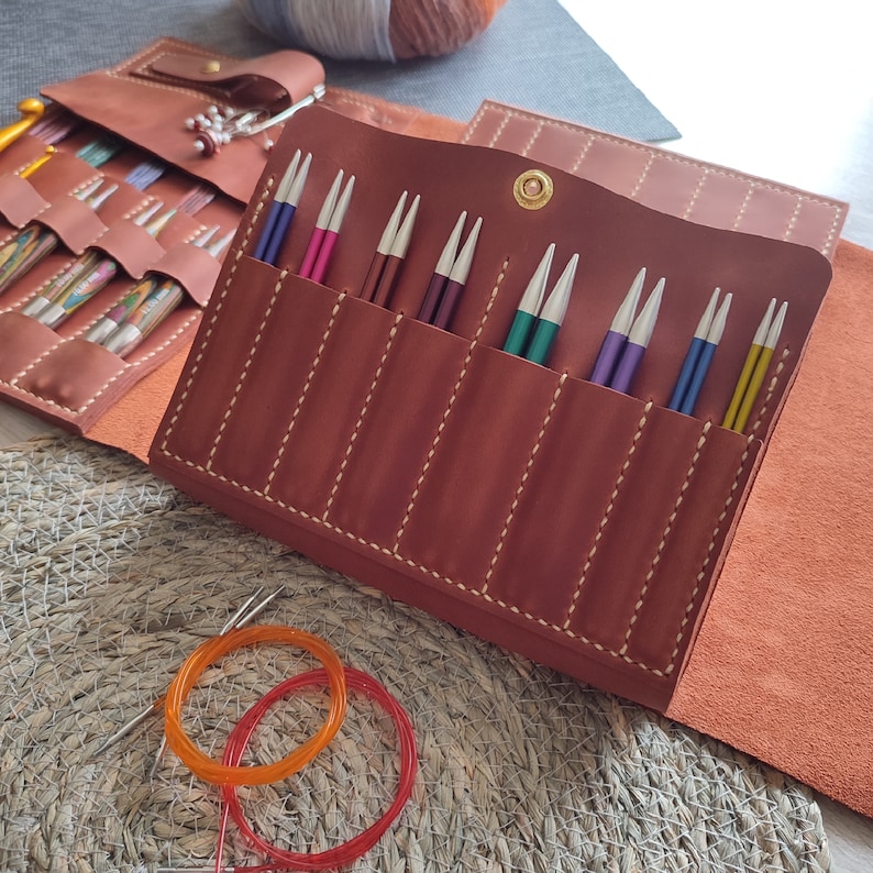 May include: A brown leather case with multiple slots for holding knitting needles. The case is open and the slots are filled with colorful knitting needles.