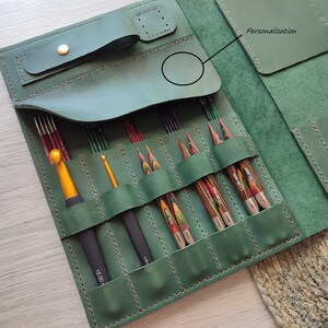 May include: A green leather case with multiple slots for holding knitting needles and crochet hooks. The case has a flap with a leather strap and a metal buckle. The case has a personalization area on the front.
