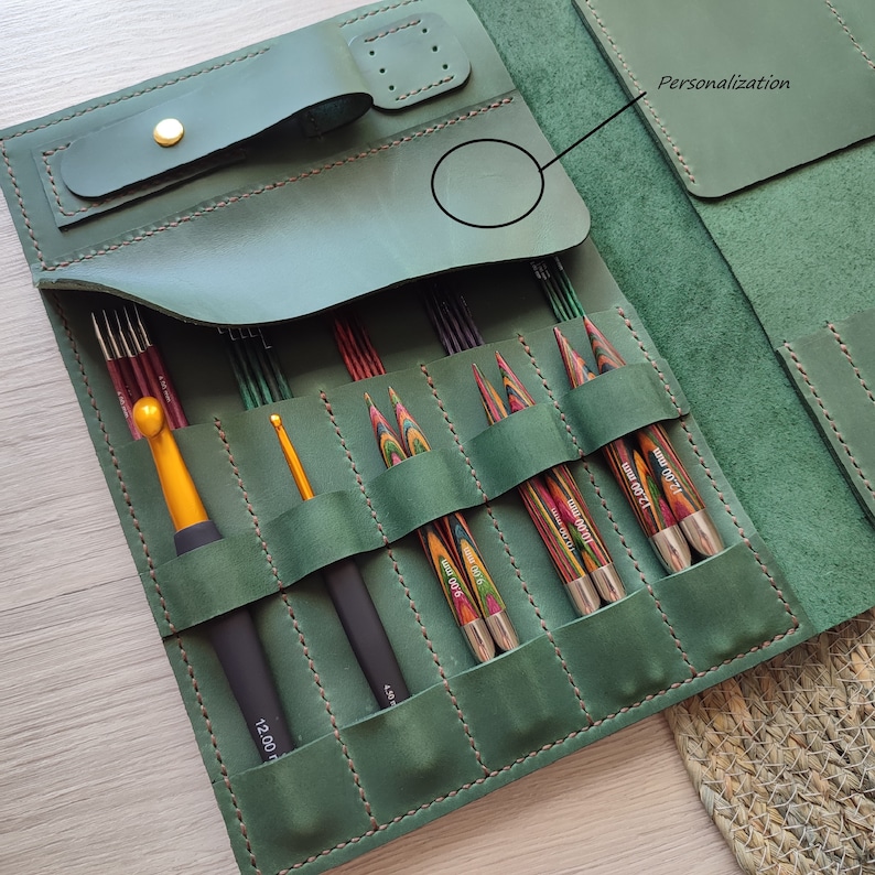 May include: A green leather case with multiple slots for holding knitting needles and crochet hooks. The case has a flap with a leather strap and a metal buckle. The case has a personalization area on the front.