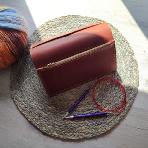 Peut inclure: Une trousse à aiguilles à tricoter en cuir brun rougeâtre avec une fermeture éclair dorée, ouverte pour révéler l'intérieur. Deux aiguilles à tricoter violettes et une aiguille circulaire rouge sont à côté de l'étui. Une pelote de laine colorée est à gauche.