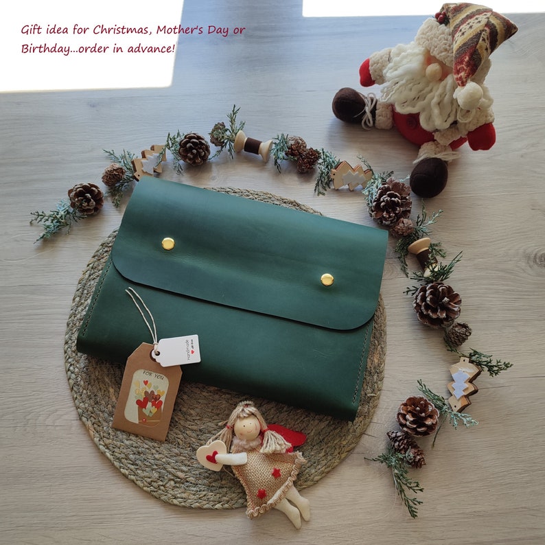 May include: A green leather clutch with gold accents and a small tag that says "For You". The clutch is on a woven mat with a small angel figurine and pine cones.