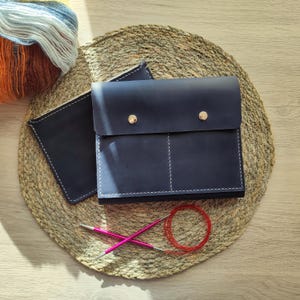 May include: A navy blue leather knitting project bag with a flap closure and two gold-colored snaps. The bag is next to a matching navy blue pouch, pink knitting needles, and a ball of colorful yarn. The items are on a woven mat.