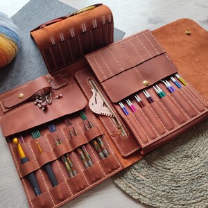 May include: A brown leather case with multiple compartments for storing knitting needles. The case is open and the needles are visible. The case has a zipper closure and a flap with a snap closure.