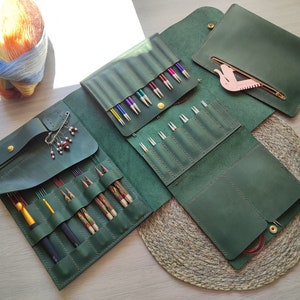 May include: A green leather knitting needle case with multiple compartments for storing needles and other accessories. The case is open to reveal a variety of needles and a pink plastic tool.