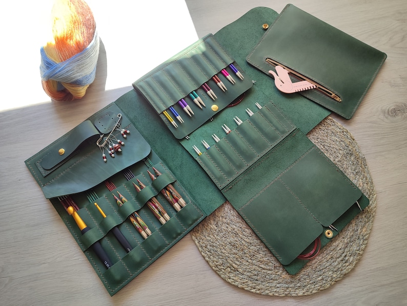 May include: A green leather knitting needle case with multiple compartments for storing needles and other accessories. The case is open to reveal a variety of needles and a pink plastic tool.