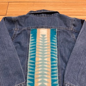 May include: A denim jacket with a rectangular patch on the back. The patch features a geometric design in shades of blue, beige, and white. The jacket has long sleeves and a classic collar.
