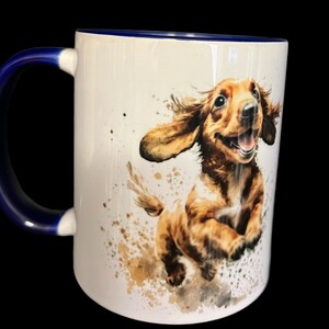 May include: White ceramic mug with a blue rim and handle. The mug features a watercolor illustration of a brown and white dachshund puppy running with its tongue out.
