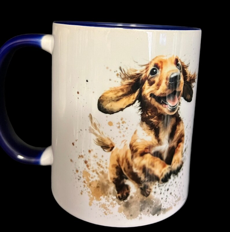 May include: White ceramic mug with a blue rim and handle. The mug features a watercolor illustration of a brown and white dachshund puppy running with its tongue out.