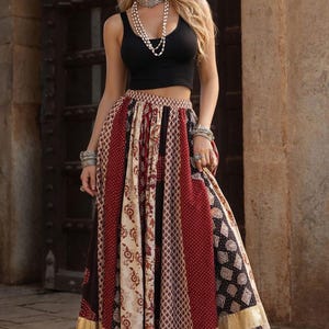 Könnte beinhalten: Ein Outfit für Damen mit einem schwarzen Crop-Top und einem langen, gemusterten Rock. Der Rock zeigt Bahnen aus roten, cremefarbenen und schwarzen Stoffen mit verschiedenen geometrischen und floralen Designs. Goldener Besatz akzentuiert den Saum. Das Outfit wird mit Silberschmuck und braunen Sandalen kombiniert.