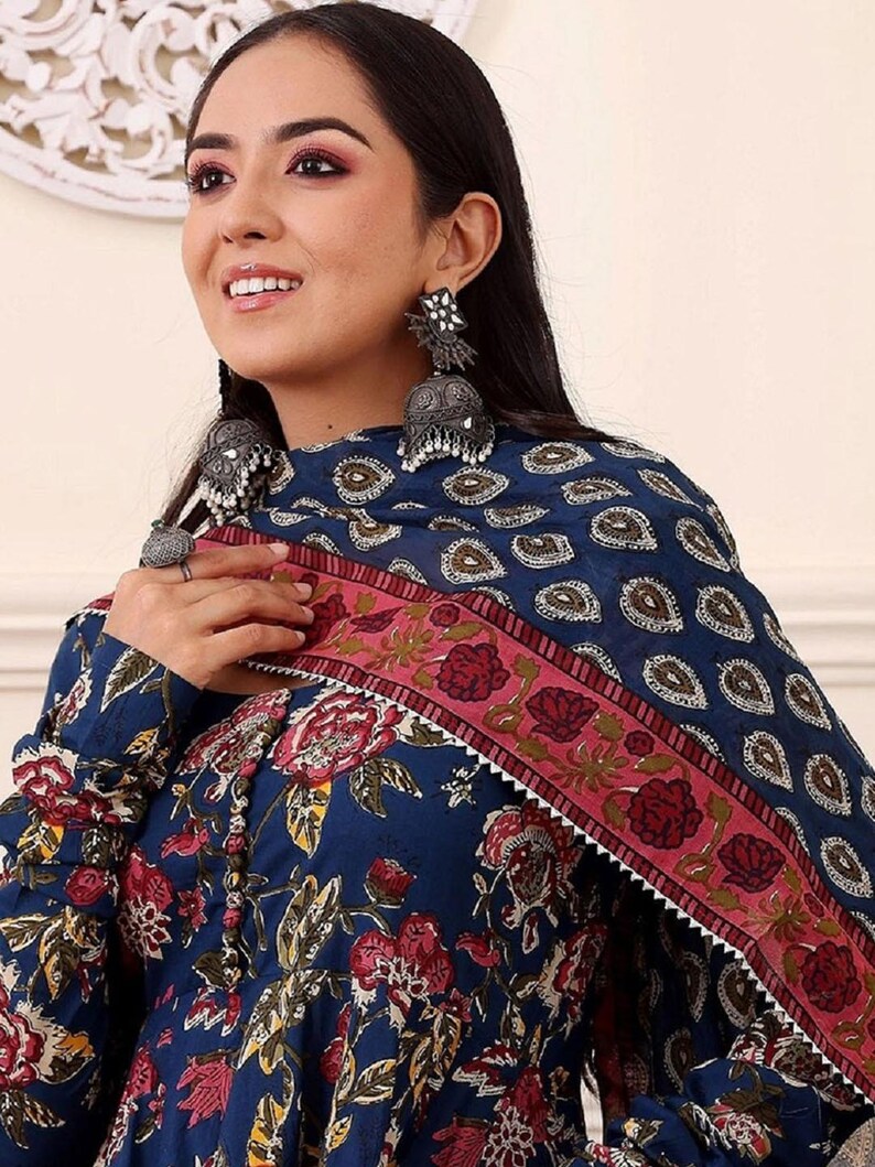 May include: A woman wearing a dark blue floral print dress with a matching scarf. The dress features a red and pink floral pattern. The scarf has a paisley design and a pink border. The woman is wearing silver earrings.