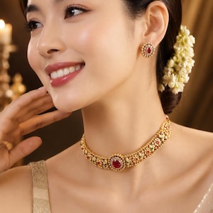 May include: A gold choker necklace and matching stud earring set featuring intricate metalwork, small white stones, and a central oval-shaped red gemstone. The necklace has a repeating floral pattern with a prominent red stone at the center.