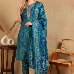 May include: A teal and blue printed salwar kameez set. The outfit includes a tunic, pants, and a matching scarf. The tunic features intricate embroidery around the neckline. The fabric has a geometric pattern.