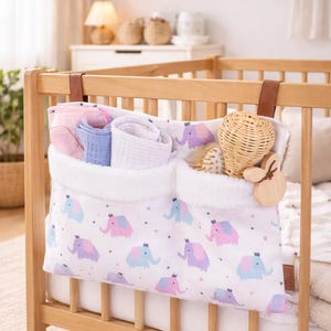 May include: A white fabric baby crib organizer with a pattern of pink, purple, and blue elephants. The organizer has two pockets with white fluffy trim and brown leather straps. The crib is made of light wood.