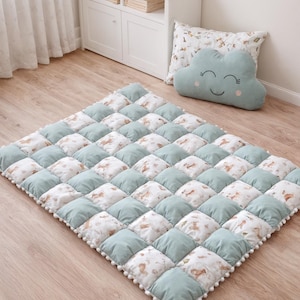 Może przedstawiać: Pikowana mata do zabawy z powtarzającym się wzorem naprzemiennych jasnoniebieskich i wzorzystych kwadratów. Wzorzyste kwadraty mają beżowo-brązowy kwiatowy wzór. Mata jest obszyta białym obramowaniem z pomponów. Widoczna jest również poduszka w kształcie chmurki i pluszowy królik.