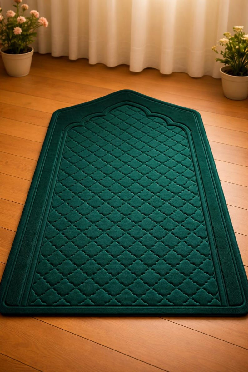 May include: A dark teal prayer rug with a quilted diamond pattern. The rug has a decorative arched top and a border. The rug is on a wooden floor.