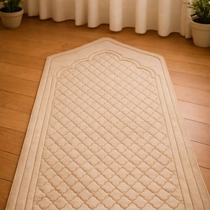 May include: Beige prayer rug with a quilted diamond pattern and an arched top. The rug is rectangular with a decorative border and is placed on a wooden floor. The background includes curtains and potted plants.