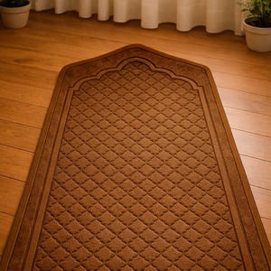May include: A brown prayer rug with a diamond pattern and an arched top. The rug is rectangular with a decorative border. The rug is placed on a wooden floor.