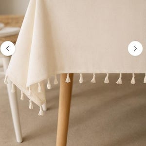 Op de afbeelding: Crèmekleurig rechthoekig tafelkleed met een gefranjerde rand. Het tafelkleed is gedrapeerd over een tafel met licht houten poten en een witte poot. De franje bestaat uit kleine kwastjes.