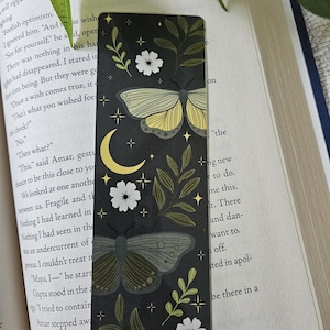 May include: A rectangular bookmark with a black background featuring two moths, a yellow crescent moon, white flowers, green leafy branches, and small four-pointed stars. The design is symmetrical and includes botanical and celestial elements.