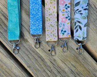 May include: Five colorful fabric keychains with silver key rings. The keychains feature different floral and geometric patterns in shades of blue, green, pink, and white.