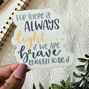 Könnte beinhalten: Ein weißer Aufkleber mit dem Text "FOR THERE IS ALWAYS light... IF WE ARE brave ENOUGH TO BE IT" in verschiedenen Farben. Der Aufkleber wird von einer Hand mit rosa Nagellack gehalten. Ein Notizbuch und ein Stift befinden sich im Hintergrund.