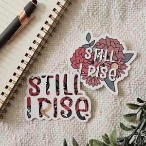 Könnte beinhalten: Zwei Blumenaufkleber mit dem Text "Still I Rise" in weißen Buchstaben. Die Aufkleber befinden sich auf einem weißen Stoffhintergrund.