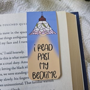 May include: A rectangular bookmark with a blue background featuring a stained glass lamp illustration at the top. Below the lamp, a yellow light beam illuminates the black text that reads, I READ PAST MY BEDTIME, in a stylized handwritten font.