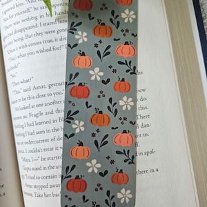 Peut inclure: Un marque-page rectangulaire sur fond vert sauge doux, orné d'un motif répétitif de citrouilles orange, de petites fleurs blanches et de tiges feuillues vert foncé. Le design est imprimé sur une surface plate aux coins arrondis.