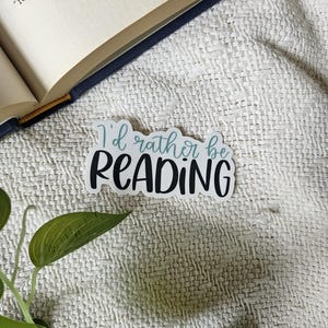 Peut inclure: Un autocollant blanc avec le texte "I'd rather be READING" en lettres noires avec un contour bleu sarcelle. L'autocollant est sur un fond en tissu blanc.