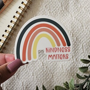Könnte beinhalten: Ein Regenbogen-Aufkleber mit dem Text "Kindness Matters" in schwarzen Buchstaben. Der Regenbogen besteht aus schwarzen, orangen, rosa, gelben und weißen Streifen.