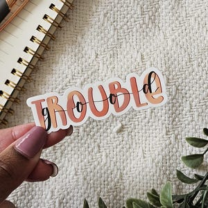 Könnte beinhalten: Ein weißer Aufkleber mit dem Text "good trouble" in orangefarbenen und rosafarbenen Buchstaben. Der Aufkleber hat eine glänzende Oberfläche und ist leicht gebogen.