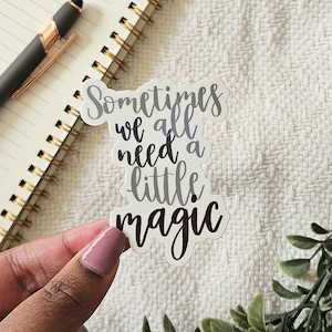 Könnte beinhalten: Ein weißer Aufkleber mit dem Text "Sometimes we all need a little magic" in einer grauen und schwarzen Schriftart.