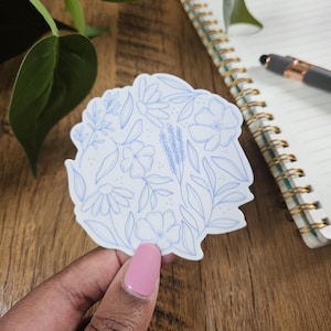 May include: A die-cut sticker featuring a light blue floral design. The sticker showcases various flowers and leaves in a delicate, line-art style. Held by a hand with a pink nail polish, the sticker is placed on a wooden surface.