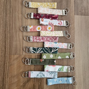May include: A set of 12 colorful fabric keychains with floral and geometric patterns. Each keychain features a silver key ring and a fabric strap with a different design.