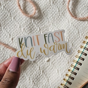 May include: A sticker with the text "Knit fast die warm" in a colorful, handwritten font. The sticker is on a white, textured surface.