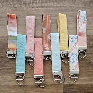 May include: Eight colorful fabric keychains with silver key rings. The keychains have different patterns and designs, including stripes, polka dots, and floral prints. Some of the keychains have inspirational quotes printed on them, such as "not be okay" and "find feel peace".