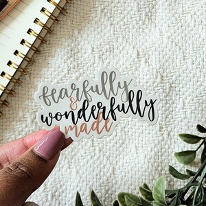 Könnte beinhalten: Ein weißer Aufkleber mit dem Text "fearfully & wonderfully made" in schwarzer und goldener Schrift. Der Aufkleber befindet sich auf einer weißen, strukturierten Oberfläche.